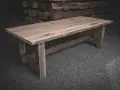 Ett rustikt bord av oljad ek med grova ben och en bred bordsskiva. Bakom syns staplade trästockar.