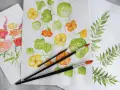 Flera akvarellmålningar med blommor och blad. En målning visar gula och orange blommor med gröna blad, en annan visar en bukett med rosa och orange blommor. Penslar ligger på pappret.