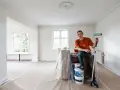 Man med färgroller och hinkar redo att måla bakom elementet i ett rum under renovering.