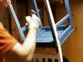 Person i orange tröja målar en blå stol med pensel.