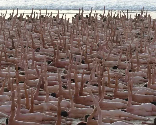 En stor grupp människor ligger på en strand, många med armarna uppsträckta. En naken målning av mänskligheten.