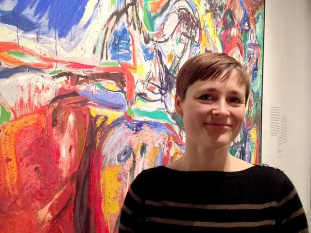 Kvinna ler framför en färgstark, abstrakt målning i Asger Jorns anda.