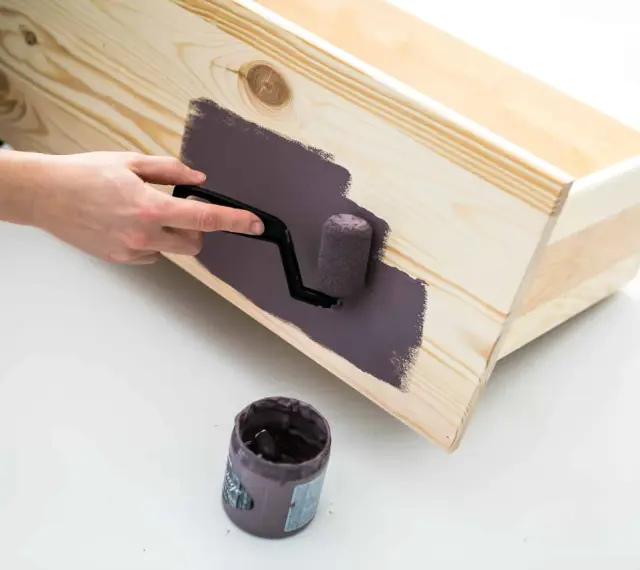 En hand använder en liten roller för att måla en trälåda med mörklila färg, vilket ger en jämn yta utan penseldrag.