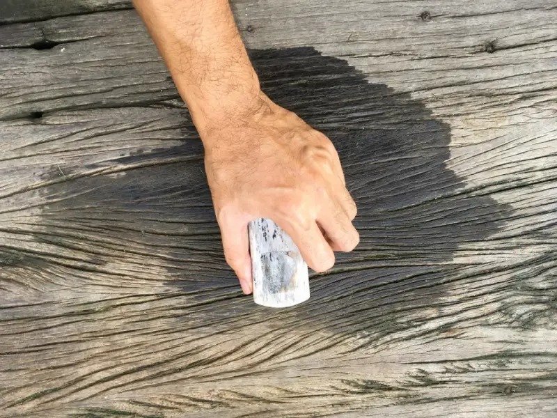 En hand håller en borste för att skura altanen. Träet är väderbitet och har en mörk, fuktig yta.